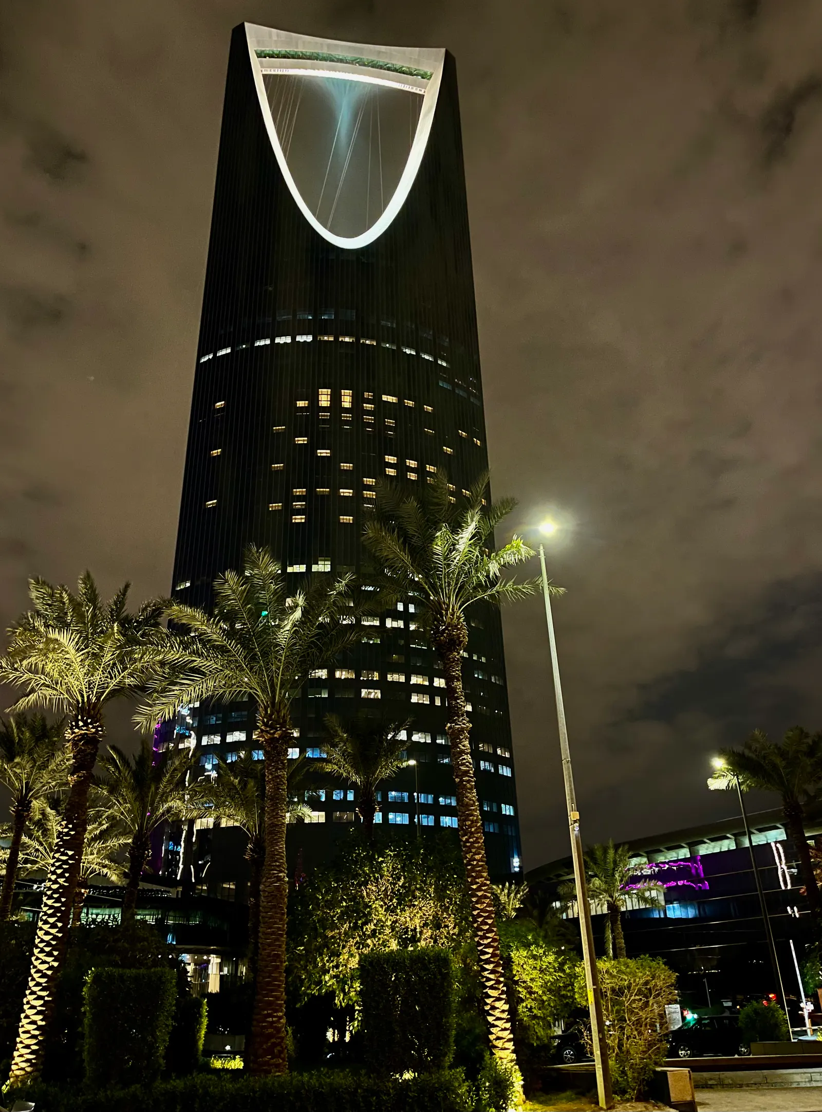 The Kingdom Centre at night, its arch outlined in white light above palm trees